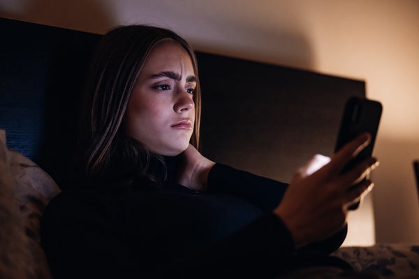 Worried looking young woman lying on her bed at night looking at her phone screen.